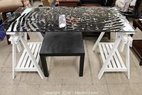 Full view of Art Deco style glass top table with many pieces of jewelry displayed on top. White wooden legs have slatted lower shelves on each side.