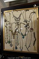 Overview photo showing assortment of over 20 necklaces laid out flat against beige fabric in display frame (not included)