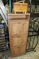Wooden cabinet with three compartments, visible shelving inside the top compartment, drop-down doors with metal handles.