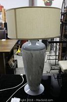 Full view of the lamp with beige fabric drum shade on black surface, showing the slender marble-effect ceramic body.