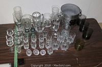 Photo showing an assortment of clear glassware including shot glasses, scotch glasses, beer mugs in clear and smoked glass, a Moet ice bucket with black label and handles, and a gold plated wine collar.