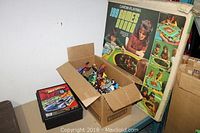 Wide shot of the boxed Carom Playing 199 Games Board, Hot Wheels carrying case, and cardboard box with action figures on a table.