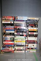 Photo showing a vertical stack of VHS tapes on pegboard background with visible movie titles including Titanic, Hot Chick, Random Hearts, Meet the Parents, Sleepless in Seattle, Austin Powers, The Way We Were, and others.