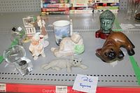 Photo showing multiple small figurines on a shelf including a reclining figure, cat, Buddha head, gnome figure, and a Greek souvenir bottle.