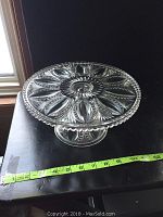Top and side view of the clear glass footed cake plate showing intricate beaded oval and fan pattern and measuring tape for size reference.