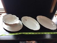 View of two small porcelain serving dishes and one gravy boat with attached drip dish sitting on a table with measuring tape showing length.