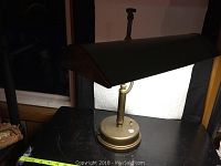 Front angled view of brass bankers lamp with green metal shade on black surface, measuring tape in front for scale.