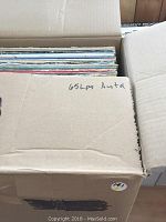 Photo of a box partially open showing a stack of LP records inside with handwritten note stating '65 LPs Asstd'.