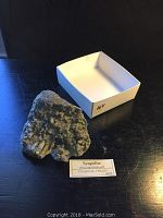 Scapolite rock sample displayed next to white collection tray and label from Craigmont, Ontario, showing crystal texture and layered rock structure.