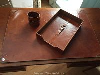 Four piece brown leather desk set showing pen holder, paper tray with buckle lid, and desk protector located on wooden desk surface.