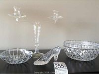 Four glass items displayed on a black surface: a large fruit bowl with cut pattern, a candy dish, a glass slipper figurine, and a tall glass candle holder.