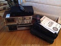 Lot of four electronic devices showing stacked VCRs, cassette player, and Nextbox with remotes and manual on top.
