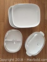 Four white ceramic baking dishes displayed on a wooden floor, two rectangular dishes without lids on top, and two oval dishes with clear glass lids below, one with an internal divider visible.