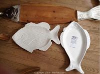 Three fish style platters: two white ceramic fish platters and one wooden fish-shaped serving plank with metal accents shown on wood floor