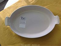 Top-down view of an oval white bakeware dish with integrated handles on each end, sitting on a yellow surface with a MaxSold sticker attached inside.