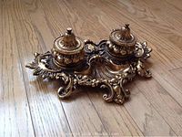 Ornate brass double ink well set sitting on wooden floor, showing detailed scrollwork and design