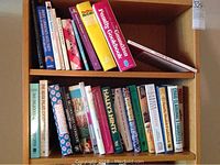 Wooden shelving unit holding multiple assorted cooking books both hardcover and paperback, different colors and sizes.