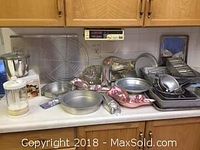 Photo showing a large assortment of metal bakeware pans including round cake pans, loaf pans, bundt pan, and various other kitchen tools and molds arranged on a kitchen counter.