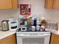Wide view of all coffee and beverage items on kitchen stove and counters including electric kettle, pitchers, mugs, and other accessories