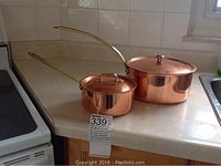 Two copper chef pots with lids, brass handles on kitchen counter near stove, showing full set and size difference.