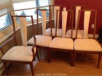 Six Peppler wood dining chairs with cream colored fabric back panels and upholstered seats arranged in two rows, on a wooden floor near windows.