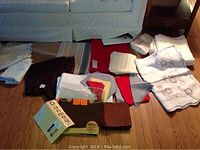 Lot of assorted tablecloths and napkins spread on the floor near a sofa, showing different colors and sizes including white embroidered and dark solid colors.