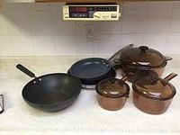 Kitchen counter showing three brown tinted Vision glass pots with glass lids and four frying pans, one Circulon deep fry pan and three other fry pans.