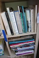 Bookshelves holding a variety of cookbooks, crafting, knitting and painting books. Titles include several by Mario Batali, Julia Child, and others focusing on Italian cooking and holiday themes. Books are a mix of hard and soft covers, some vintage with wear.