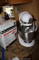 Photo of KitchenAid Classic mixer unit with stainless steel bowl, placed on countertop, and boxed food grinder attachment next to it.