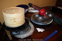 Three-tier bamboo steamers, a pewter southwestern platter, metal roasting pan, hand painted mixing bowls, cupcake baking pans, and a red manual citrus juicer all stacked on table shown.