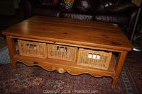 Front view of the oak coffee table showing rectangular shape, carved detail on bottom panel, and three wicker baskets in storage compartments.
