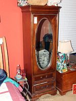 Full view of a tall wooden armoire with an oval mirror in the center, two drawers underneath, and placed next to a nightstand with a table lamp.