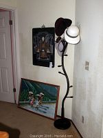 View of the Asian lacquer framed art mounted on wall, Haitian painting leaning against wall, and metal hat rack with hats.