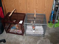 Two metal bird cages side by side, one dark red and one gray, both with toys and food bowls inside, placed on a floor against a wooden wall.