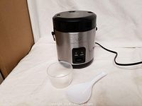 Full view of the Wolfgang Puck rice cooker including attached power cord, plastic measuring cup, and white plastic rice paddle on a white cloth background.