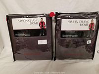 Two sealed Simon Chang Home window panel packages side by side, dark color fabric visible through packaging.