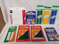 Overview showing all 9 pieces including Velcro sticky back tape, Fantastick tacky squares, mounting squares in orange and red packaging, and three fastening tapes in green, yellow, and blue packaging.