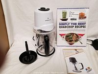 Wolfgang Puck quad chopper with stacking blades, recipe book, and instruction manual on a white fabric background.