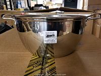 Front view of a shiny stainless steel pot with two handles and a label on the side, placed on a cardboard box.