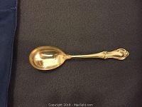 Cream soup spoon placed flat on dark fabric backing to show full front face, illustrating ornate handle and bowl shape.
