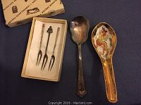 Photo of all five pieces laid out on a dark blue background: three small silver forks in a box, a silver spoon, and a ceramic soup spoon with a dragon design.