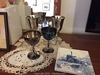 Four silverplate goblets with reflective surfaces on crocheted doilies next to ceramic tile with windmill pattern.