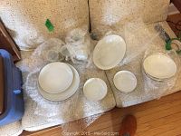 Variety of white and gold rimmed plates, bowls, cups, and serving dish displayed laid out on a couch surface with bubble wrap.