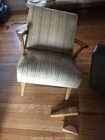 Front view of mid century modern upholstered chair showing greenish beige striped fabric and detached broken wooden leg pieces on floor.
