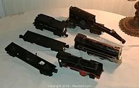 Seven vintage model train parts on table, including locomotives and flat cars, dark colors with some paint wear.