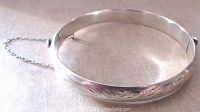 Front angle view showing engraved floral/leaf detail and safety chain attached to sterling silver bangle bracelet.