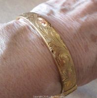 Close-up view of bracelet worn on wrist showing detailed scrolled engravings on the gold tone surface.