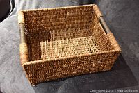 Photo showing a rectangular wicker basket with wooden handles on a dark fabric surface.