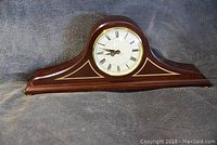 Front view of wooden mantle clock showing round white clock face with black Roman numerals and gold-tone trim on dark wood body.