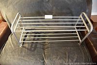 Front angled view of lightweight silver metal shoe rack with two shelves of parallel metal rods, placed on a dark fabric surface.